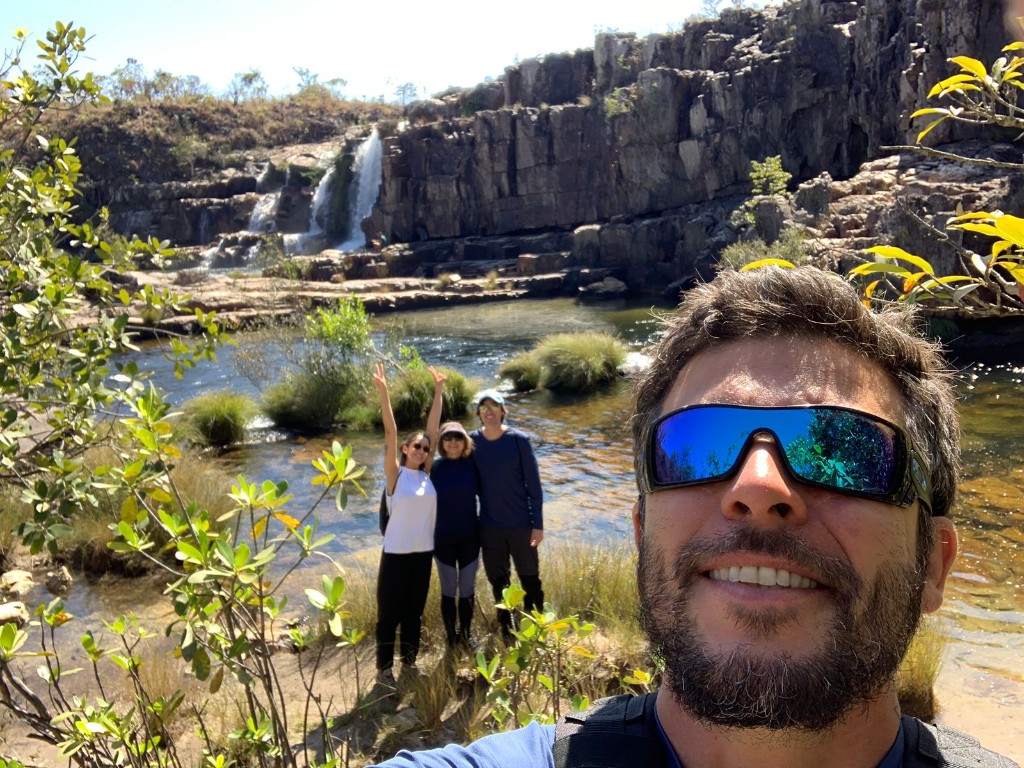 Guia e turistas em selfie à beira de rio com cachoeira ao fundo na Chapada dos Veadeiros