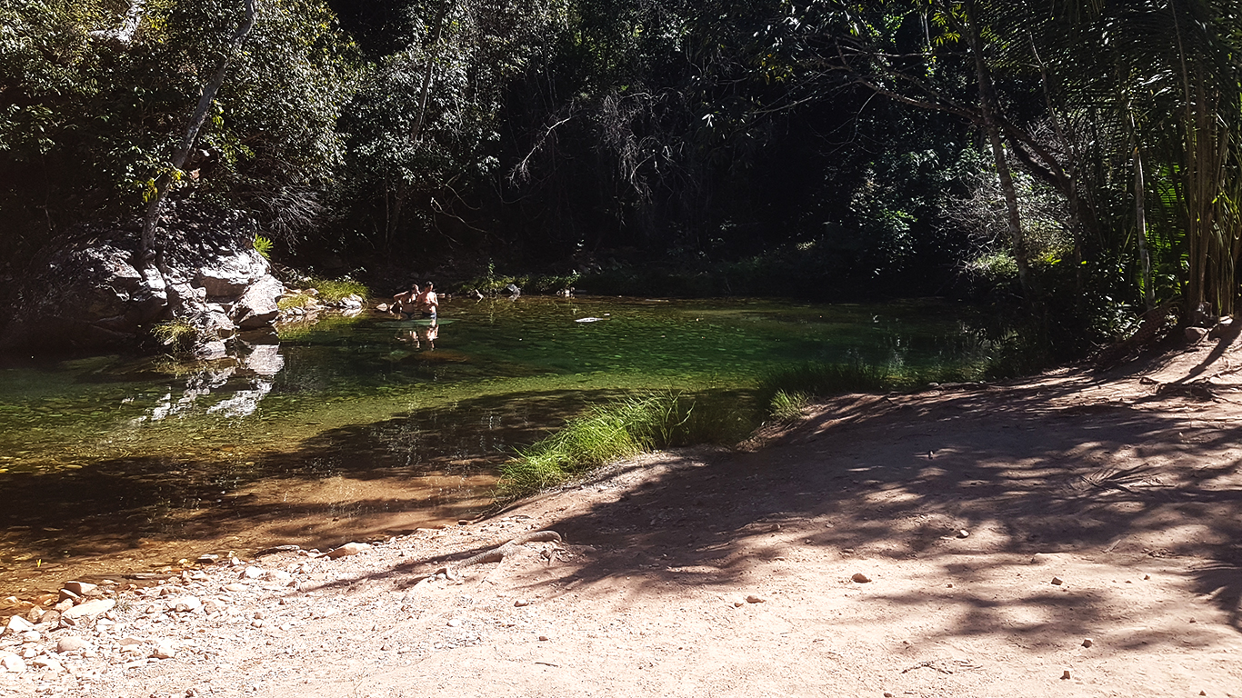 Poço verde esmeralda da Cachoeira do Segredo em São Jorge na Chapada dos Veadeiros, com reflexo das árvores na água calma e trilha de areia na margem