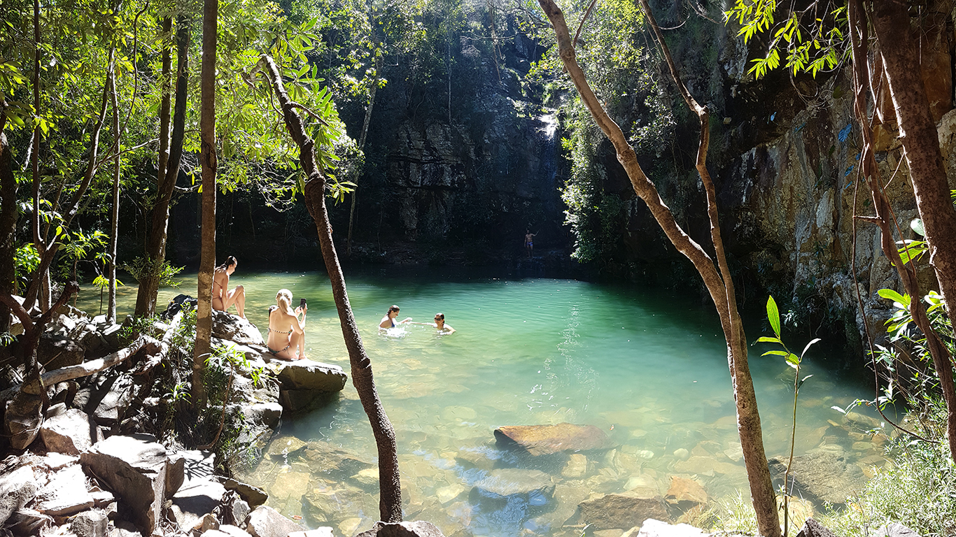 Turistas no Poço do Sol da Cachoeira Loquinhas na Chapada dos Veadeiros durante o período de chuvas, com água esverdeada entre rochas e mata fechada